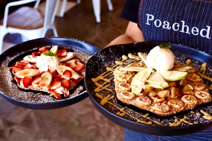 A member of the Poached staff team holding a plate of strawberry and banana waffles with a scoop of ice cream in their right hand and some apple pie waffles with a scoop of ice cream in their left hand at Poached Breakfast Cafe, Chalong, Phuket, Thailand - one of the best cute brunch places in Phuket