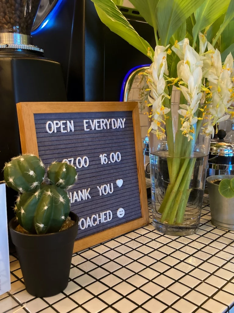 Small black sign saying "Open everyday 7am-4pm, Thank You, Poached". It is surrounded by plants and coffee paraphernalia at the best cute coffee shop in Chalong: Poached Breakfast Cafe, Chalong, Phuket, Thailand