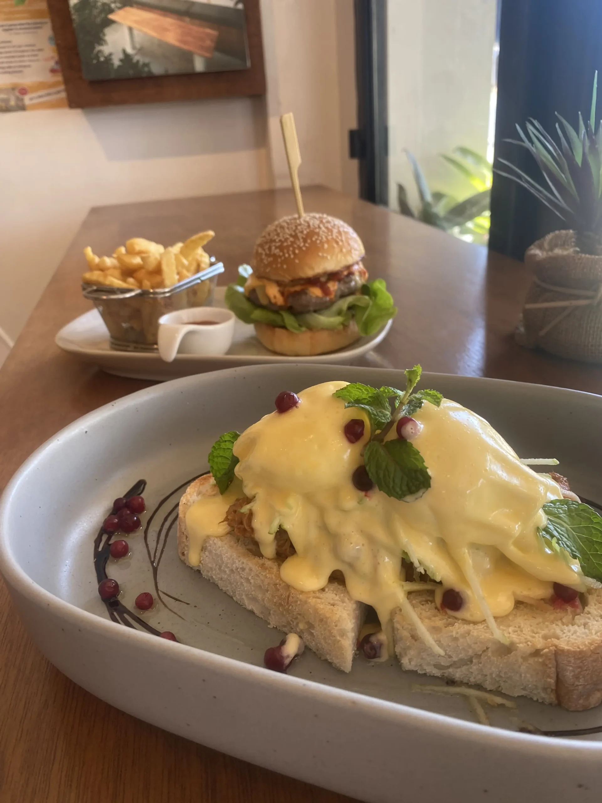 A plate with pulled pork eggs benedict topped with fresh mint and there is a plate in the background with a delicious looking stacked burger and fries. The perfect accompaniment to an iced coffee near Chalong. Only at Poached Breakfast Cafe, Chalong, Phuket, Thailand
