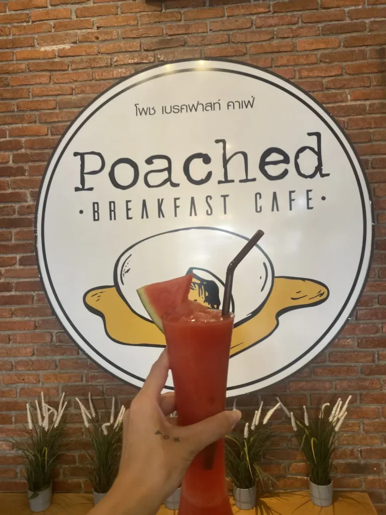 Someone holding a watermelon smoothie in a glass with a straw and slice of watermelon in front of the Poached Breakfast Cafe sign. A delicious companion to a fresh salad in Chalong that can only be found at Poached Breakfast Cafe, Chalong, Phuket, Thailand