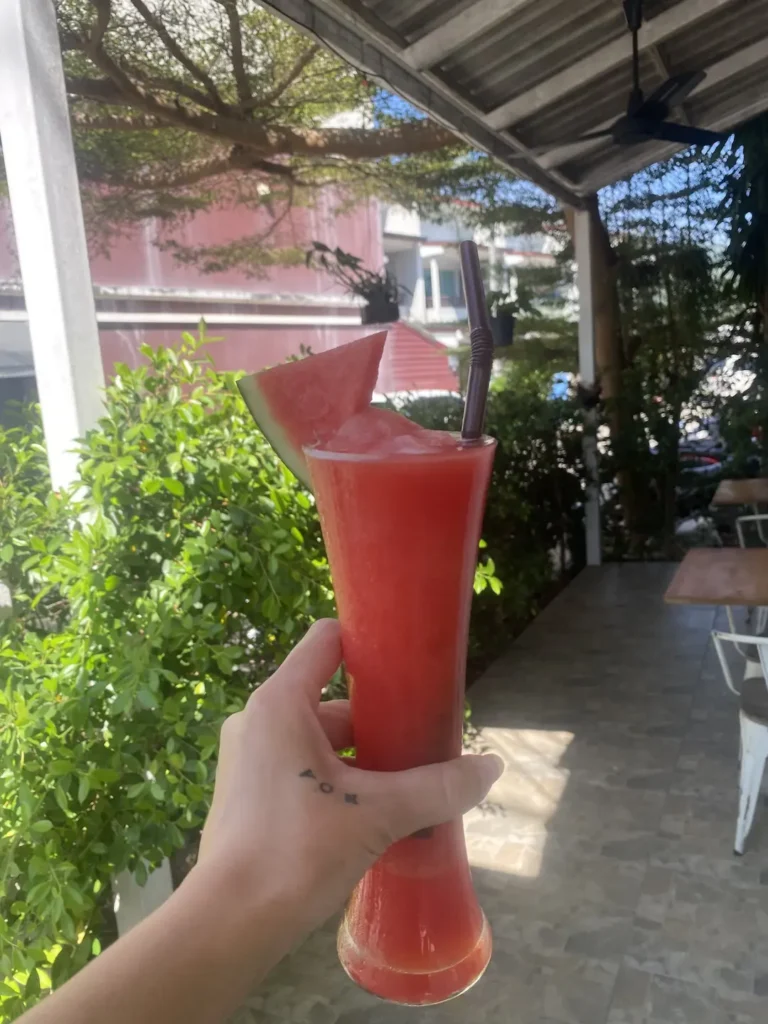 A hand holding up a cold watermelon smoothie in a small garden area under the cover of some shade. The perfect alternative to an iced coffee near Chalong. Only at Poached Breakfast Cafe, Chalong, Phuket, Thailand