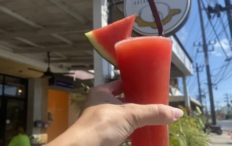 Watermelon smoothie in the sunshine being held up to our sign by a hand. A great alternative to street food in Chalong. Only at Poached Breakfast Cafe, Chalong, Phuket, Thailand