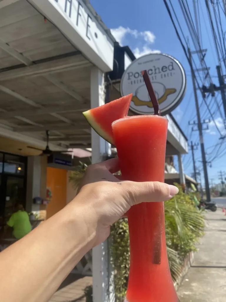 Watermelon smoothie in the sunshine being held up to our sign by a hand. A great alternative to street food in Chalong. Only at Poached Breakfast Cafe, Chalong, Phuket, Thailand