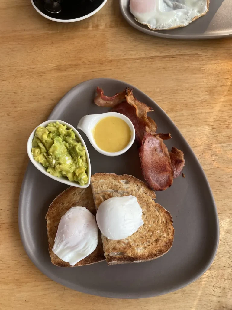 Smashed avo, 2 poached eggs, sourdough bread, bacon and hollandaise sauce on a plate. The best meal to eat before a day of island hopping in Phuket.