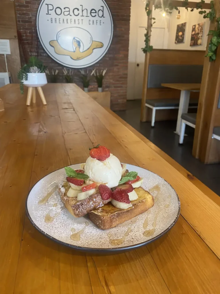 A plate of french toast with banana, ice cream and strawberries, drizzled with syrup and placed on a long wooden table at the best place to fuel up before a day of snorkelling in Phuket - Poached Breakfast Cafe, Chalong, Phuket, Thailand